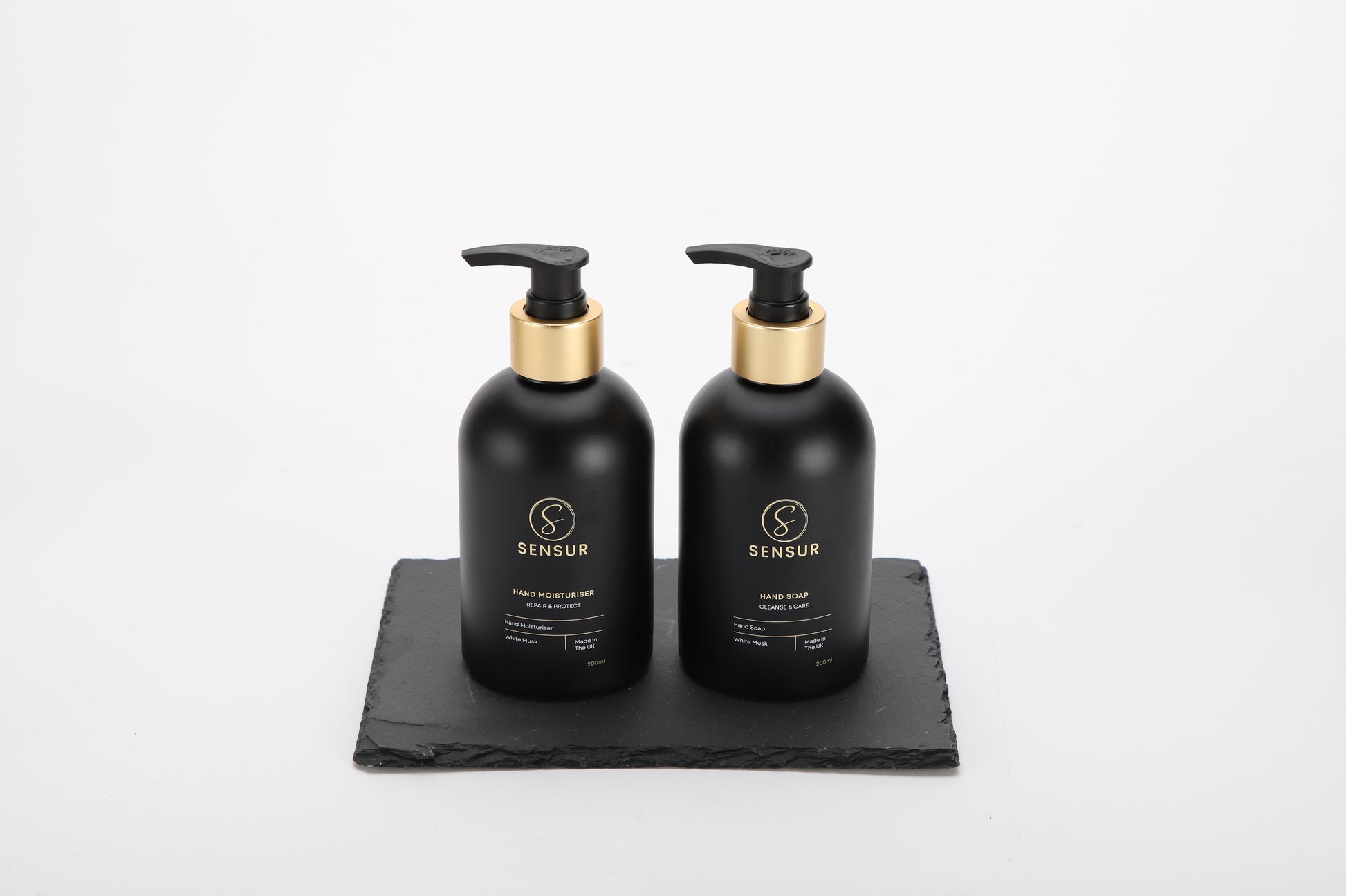 Two black bottles with gold pumps on a black slate tray against a white background