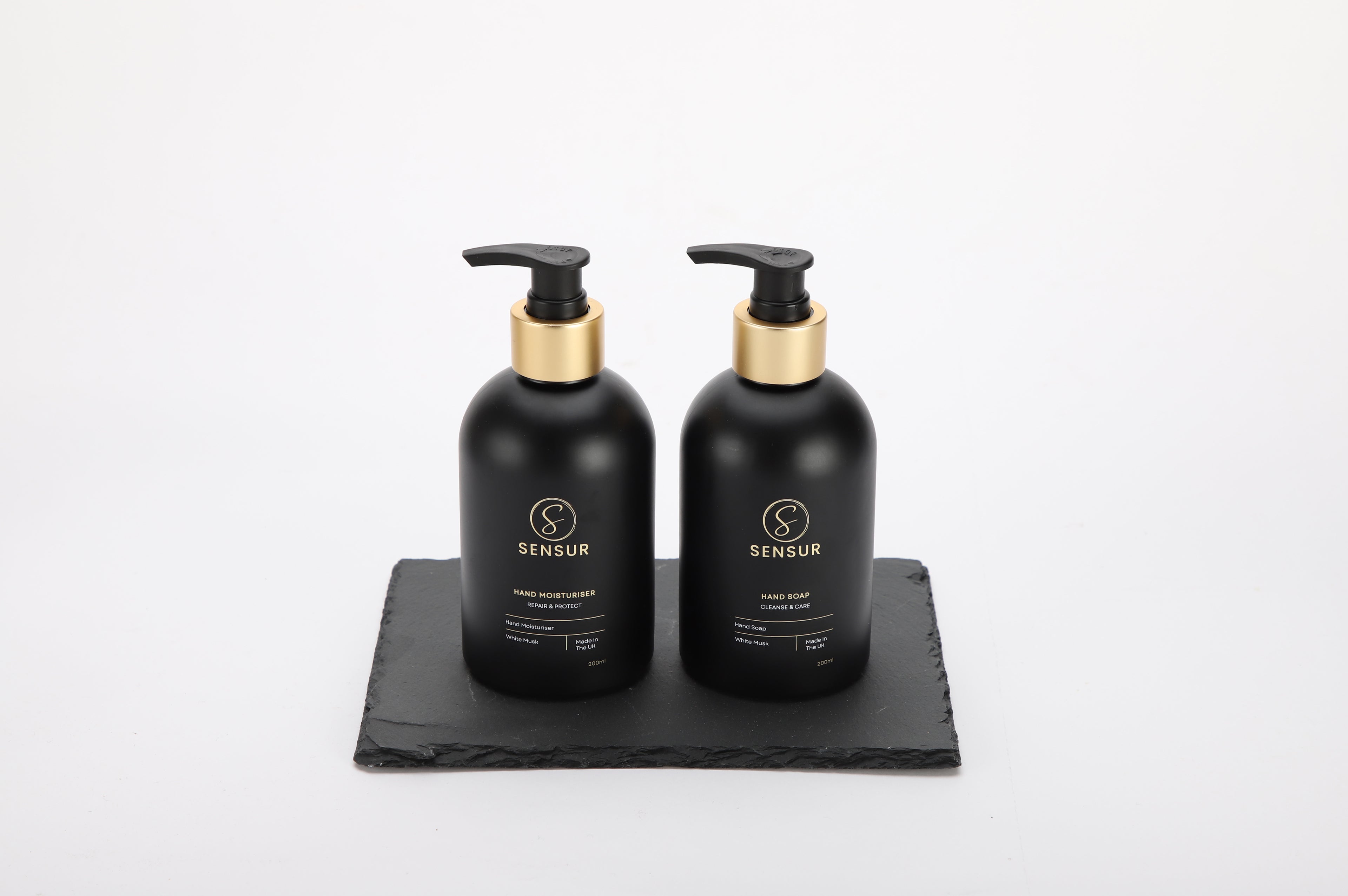 Two black bottles with gold pumps on a black slate tray against a white background